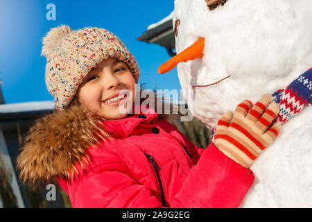 Vacanza invernale. Ragazza in piedi all'aperto vicino casa rendendo pupazzo di neve il naso con la carota sorridendo felice close-up Foto Stock