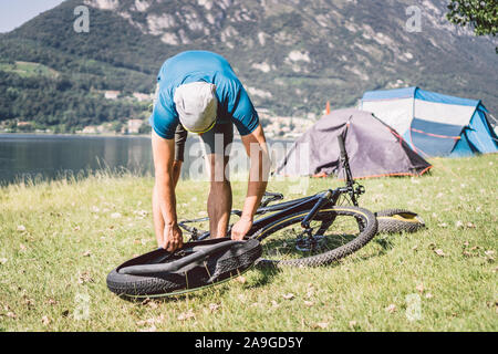 La riparazione di biciclette. Uomo che ripara la Mountain Bike. Ciclista uomo in difficoltà posteriore ruota ruota caso di incidente. Correzioni uomo Bike nei pressi del lago in Italia lo sfondo Foto Stock