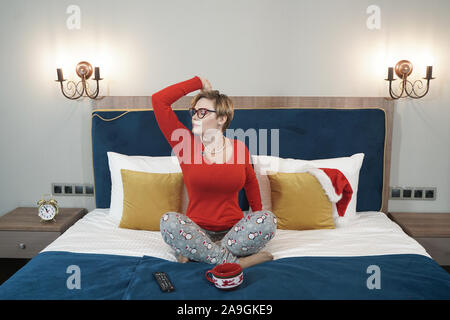 Carino plus taglia i capelli corti ragazza bionda in bicchieri sbadigli al mattino nel letto seduto in pigiama e stretching. Foto Stock