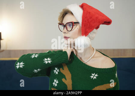 Carino dimensione plus nerd ragazza in bicchieri e verde maglione di Natale con le renne e rosso di Santa Claus hat al mattino sul letto in posa. Foto Stock
