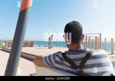 Vista posteriore di un uomo seduto sulla promenade guardando all'orizzonte Foto Stock