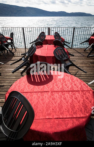 Sedie nere sono ribaltati contro l'esterno cafe tabelle con colore rosso brillante tovaglie sui decking accanto al Lago di Garda Foto Stock
