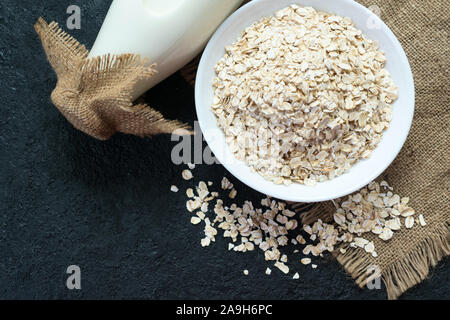 Una bottiglia di latte di avena e una piastra di fiocchi d'avena su uno sfondo nero. Il concetto di cibo vegetariano. Foto Stock