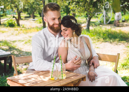 Coppia attraente sposi novelli, felice e gioioso momento. sposa e lo sposo sedersi a tavola insieme per due nel bosco. Concetto romantico data. Coppie in viaggio di nozze seduta Foto Stock