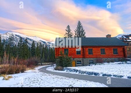 Sentiero innevato a piedi con rustica cabina in legno presso il Canmore Spring Creek Alpine Mountain Village, con i suggestivi colori del Sunset Sky sfondo Foto Stock