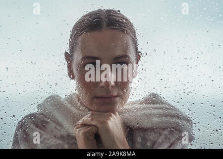 Ritratto di una donna triste guardando dall'esterno nella casa attraverso una finestra coperta con gocce di pioggia, autentico ritratto di una ragazza calma Foto Stock