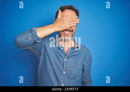 Bella la mezza età senior uomo con i capelli grigi isolati su sfondo blu a sorridere e ridere con la mano sulla faccia che copre gli occhi per la sorpresa. C cieco Foto Stock