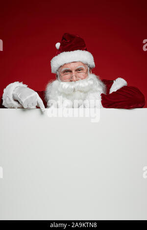 Ritratto di Babbo Natale con la barba bianca guardando la fotocamera e puntando al tabellone vuoto isolato su sfondo rosso Foto Stock