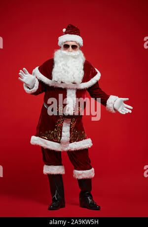 Ritratto di Babbo Natale in rosso splendido costume indossando occhiali da sole e balli contro lo sfondo rosso Foto Stock