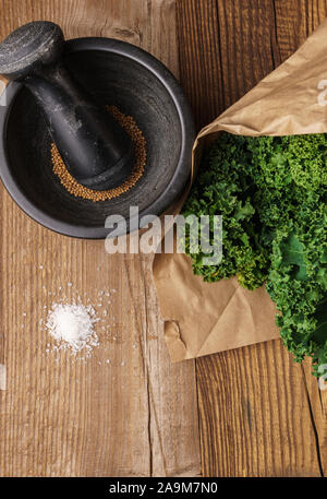 Disposizione di kale in sacchetto di carta, nero mortaio e pestello con semi di senape, un mucchio di sale marino grosso a trama sulla superficie di legno - dall'alto verso il basso Foto Stock