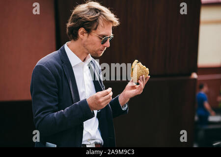 Il successo e il bel giovane in vestiti dell'Ufficio di mangiare un panino sulla strada a pranzo Foto Stock