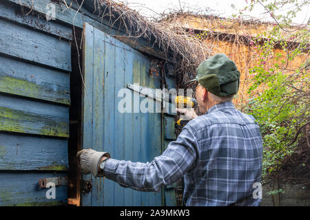 L'uomo la demolizione di un vecchio giardino in legno passo - utilizzando un cacciavite elettrico per rimuovere le cerniere della porta Foto Stock