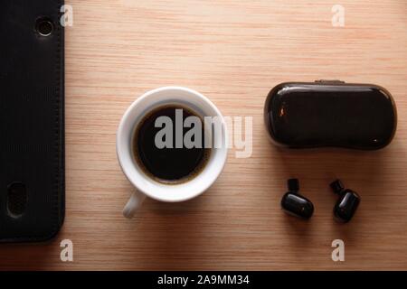 Gli auricolari Bluetooth accanto ad un mobile e una tazza di caffè su una scrivania in legno. Disposizione piatta con copia vuota dello spazio. Foto Stock