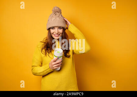 Una bruna è in piedi su uno sfondo giallo in un maglione giallo con un Thermo Mug, guardando la telecamera, regolando il suo cappello Foto Stock