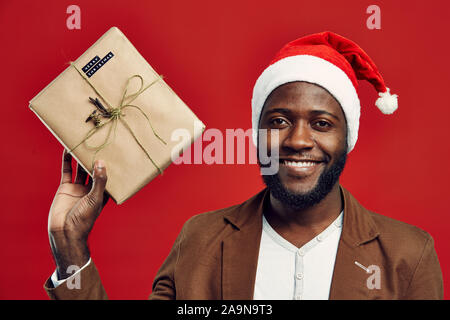 Vita ritratto di felice afro-americano di uomo che indossa Santa hat e tenendo presente mentre posa contro lo sfondo rosso Foto Stock
