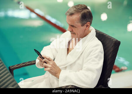 Angolo di alta vista sul moderno uomo senior indossare accappatoio utilizza lo smartphone durante i momenti di relax a luxury spa resort, spazio di copia Foto Stock