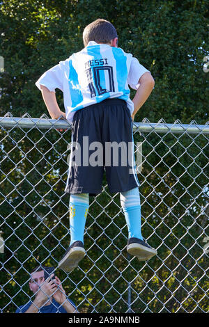 Giovane ragazzo indossa Messi maglia jersey n. 10 e salendo una catena collegamento recinto metallico in un parco in Castro Valley, California. Foto Stock