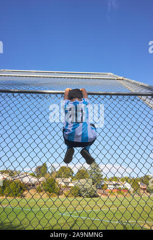 Giovane ragazzo indossa Messi maglia jersey n. 10 e salendo una catena collegamento recinto metallico in un parco in Castro Valley, California. Foto Stock