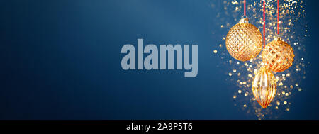 Tre golden baubles di Natale appeso contro lo sfondo blu scuro con polvere d'oro. Anno nuovo banner. Foto Stock