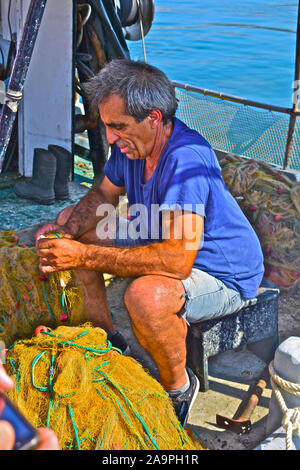 Un tipico greco fisherman rammendo / Pulizia reti a bordo la sua piccola barca da pesca ormeggiate presso la banchina a Argostoli. Foto Stock