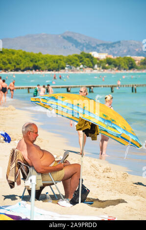 Alcudia, Spagna 14.09.2011 - Vecchio Uomo sovrappeso leggendo un giornale in spiaggia di sabbia sotto l'ombrello. La gente a prendere il sole a Playa de Muro. Mallorca Foto Stock