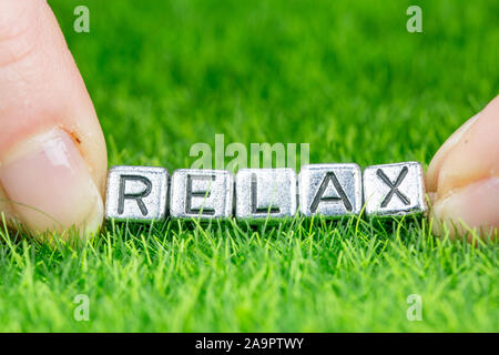 Chiudere fino sulla parola RELAX scritto in lettere di metallo prevista sull'erba e trattenuto tra le dita di una donna. Concetto di sfondo wellness Foto Stock