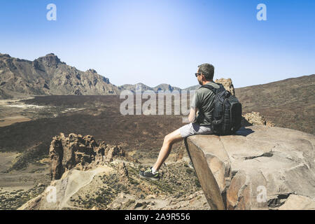 Uomo sportivo con zaino e gode di una bellissima giornata dopo la scalata da solo. Ragazzo caucasico guardando lontano seduto su una montagna scogliera affacciata sul panorama Foto Stock