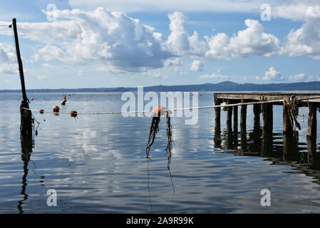 Pontile in legno per le barche contro un cielo blu con alcune nuvole. Boe galleggianti e le reti da pesca. Colline sullo sfondo. Il lago di Bolsena, Italia. Foto Stock