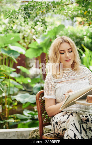 Positivo piuttosto giovane donna caucasica disegno in sketchbook quando in appoggio tra la lussureggiante vegetazione Foto Stock