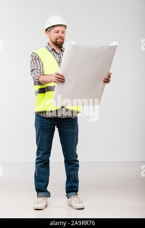 Felice giovane barbuto builder in bianco casco e abbigliamento da lavoro guardando il blueprint Foto Stock