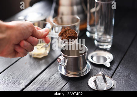 Preraring gocciolare caffè in stile vietnamita. Man mano versando il caffè macinato in metallo caffettiera phin su sfondo nero in primo piano Foto Stock