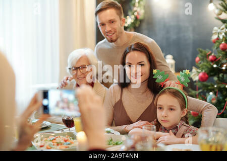 La famiglia felice seduta servita dalla mensa conviviale e guardando la fotocamera dello smartphone Foto Stock