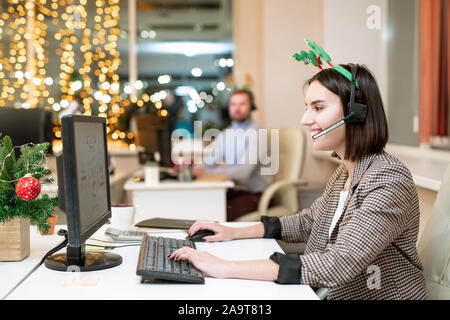 Felice giovane operatore in xmas archetto auricolare e lo shopping online dal posto di lavoro Foto Stock