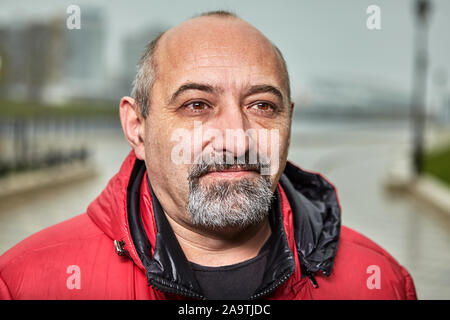 Ritratto di strada di un paffuto balding uomo con una barba grigia. Di mezza età imprenditore fotografato vicino a nuvoloso meteo, giornata d'autunno. Foto Stock