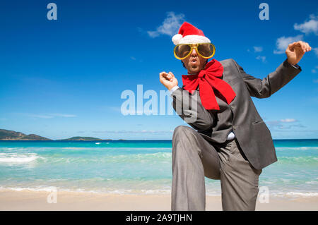 Happy businessman indossando Santa cappello, occhiali da sole e rosso grande prua di Natale danza su una spiaggia tropicale Foto Stock