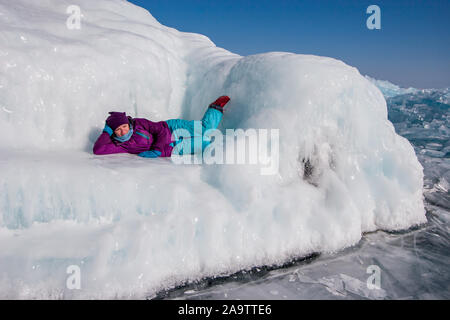 Una ragazza in abbigliamento invernale si trova su un enorme divano di ghiaccio in una giornata di sole sul lago Baikal. Ghiaccio liscio brilla al sole e fusa. Foto Stock