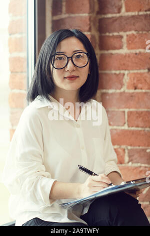 Ritratto di successo imprenditrice asiatici che indossano occhiali guardando la fotocamera mentre la scrittura di note seduta dalla finestra di office Foto Stock