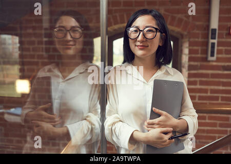 Vita Ritratto di giovane imprenditrice asiatici che indossano occhiali sorridente alla telecamera mentre appoggiata contro la parete di vetro con la riflessione, spazio di copia Foto Stock
