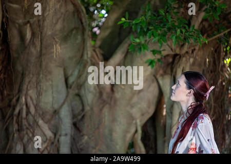 Piuttosto dai capelli rossi ragazza guardando la scala di un grande vecchio albero di fico in un parco Foto Stock