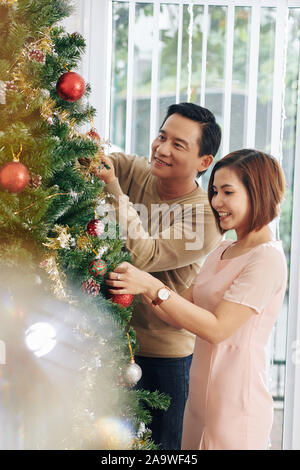 Allegro di mezza età giovane vietnamita godendo la decorazione di albero di Natale con baubles insieme Foto Stock