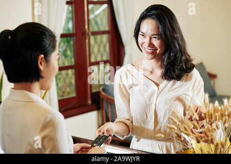 Bella sorridente giovane donna asiatica dando carta di credito alla reception per pagare il trattamento in spa salone Foto Stock