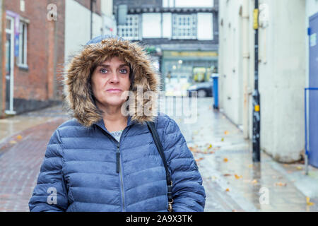 Una donna di mezza età è in piedi per strada. Indossa un impermeabile con il suo cappuccio mentre piove. Foto Stock