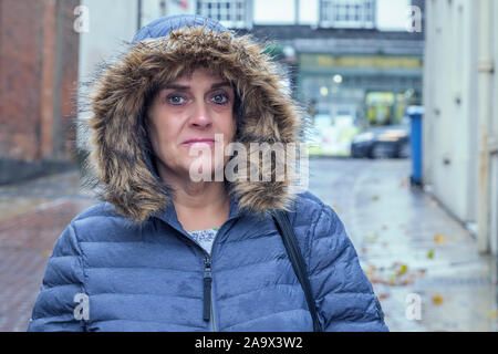 Una donna di mezza età è in piedi per strada. Indossa un impermeabile con il suo cappuccio mentre piove. Foto Stock