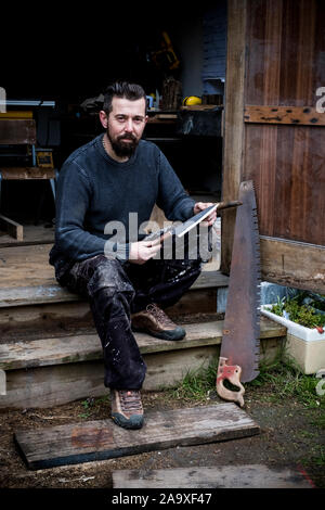 Uomo Barbuto seduto su passi al di fuori della sua officina, azienda artigianale di coltello, guardando la fotocamera. Foto Stock