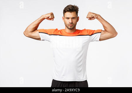 Sassy forte e bel giovane atleta, avendo cura di corpo, indossando activewear, sport t-shirt, che mostra i muscoli, alzare le mani e bicipiti brag Foto Stock