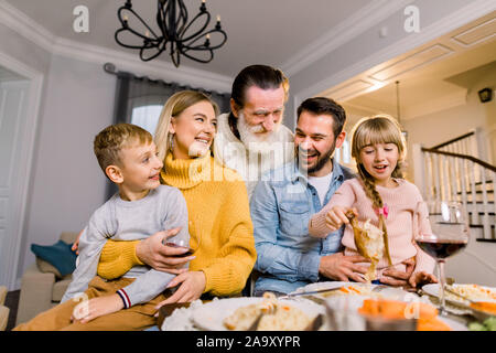 Bella serata, incontro di famiglia, sale riunioni. Allegro grey-haired nonno, i nipoti, i genitori seduti a tavola, mangiare gustoso cibo squisito e avente Foto Stock