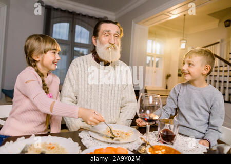 Vecchio bello nonno con i suoi due nipoti seduti al tavolo della cucina e mangiare la pasta. Bambina e ragazzo nonno di alimentazione con pasta Foto Stock