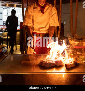 La cottura della carne di manzo ricetta teppanyaki Giapponese grigliate di carni bovine tomahawk bistecca nel recipiente di cottura utilizzando il raschiatore con fiamma in background Foto Stock
