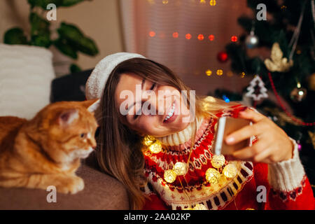 Celebrazione della Messa di Natale con cat. Donna prendendo selfie con PET in Santa's hat dal nuovo anno albero a casa. Foto Stock