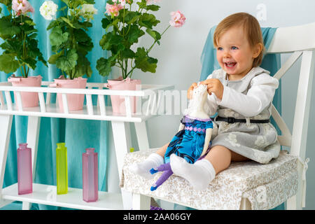 La bambina è seduta su una grande poltrona e sorridente. Ragazza con una bambola Foto Stock